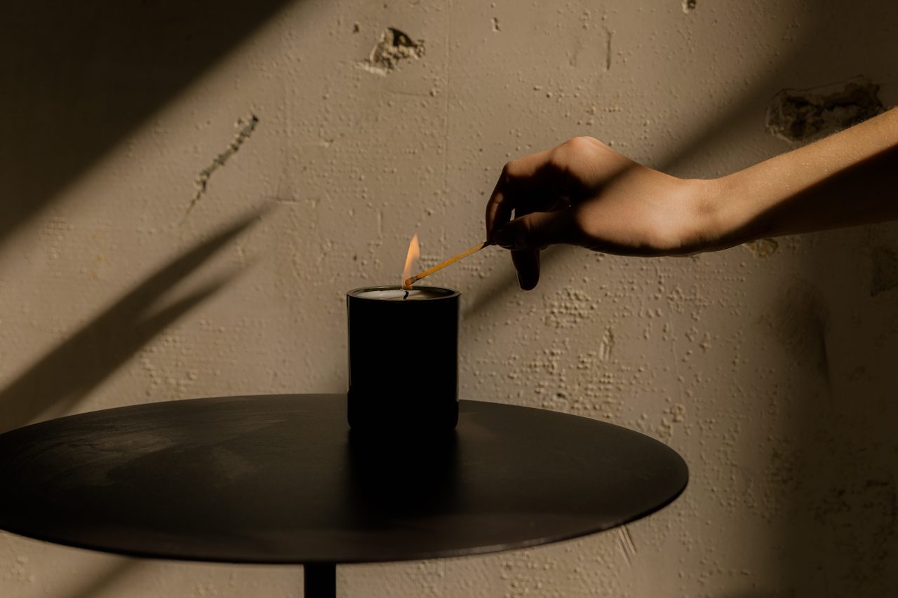 A hand lighting a black candle on a small round table against a textured plaster wall