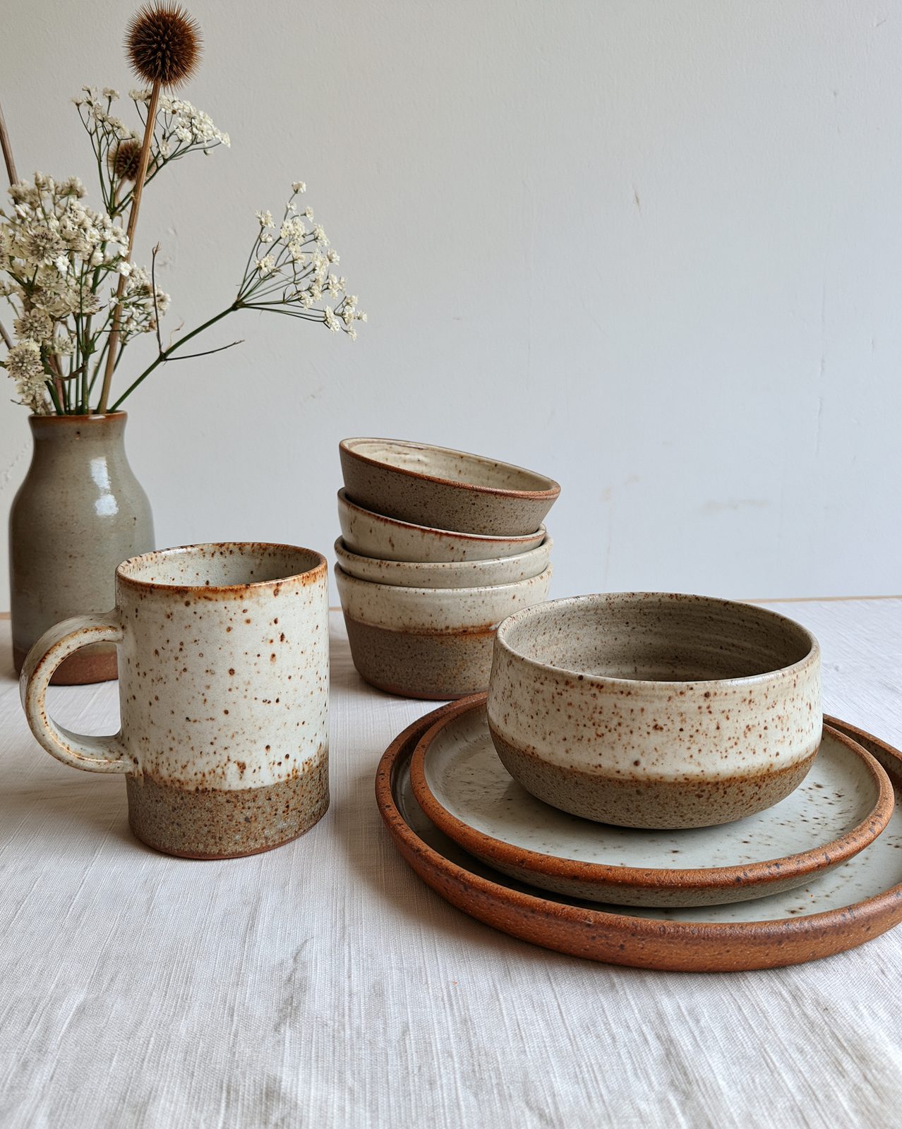 A speckled stoneware mug, two bowls, and a small vase of dried flowers on a plain table, soft daylight