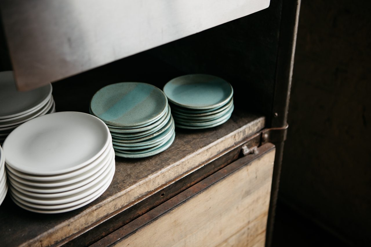 Stacks of handmade plates, white and teal, on a worn wooden shelf in a studio